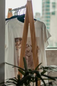 Stylish white t-shirt with art print hanging indoors on a wooden rack surrounded by casual wear.
