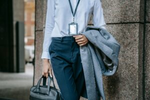 Crop faceless female entrepreneur in stylish formal outfit and name tag on neck walking on city street with briefcase and coat in hands